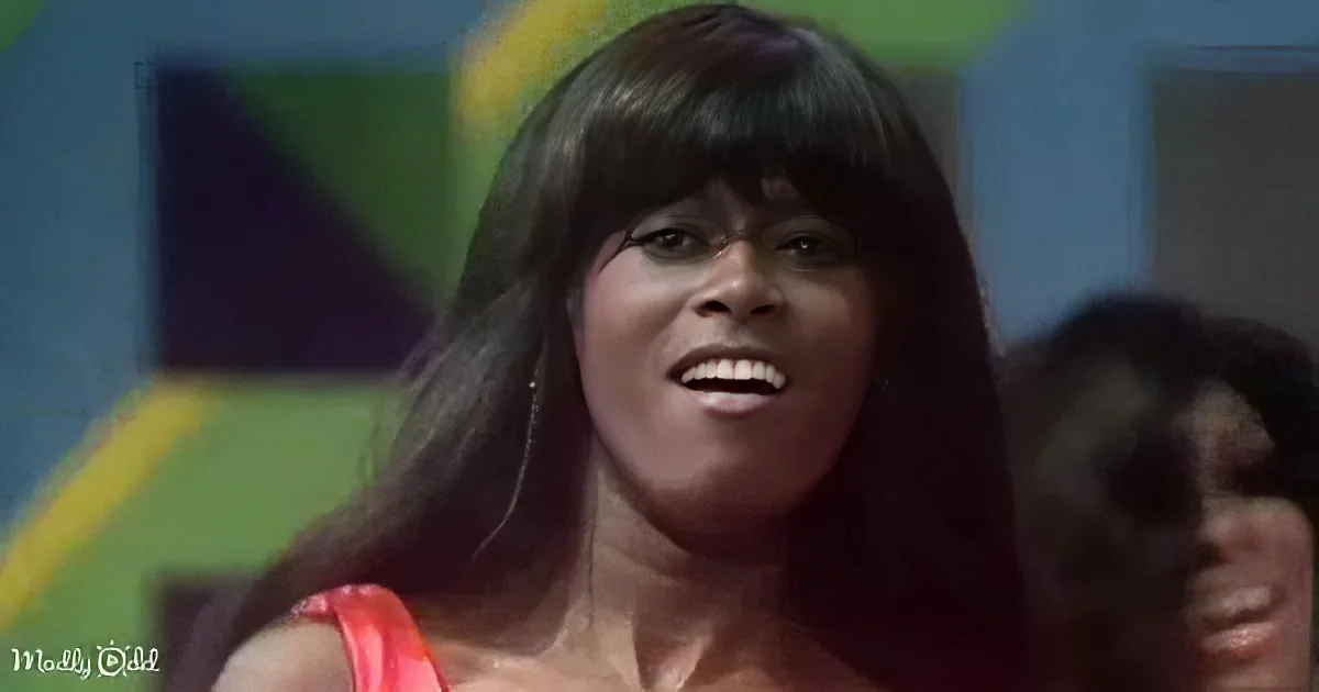 A woman with long dark hair and a red top smiles, with a colorful background and another person partially visible.