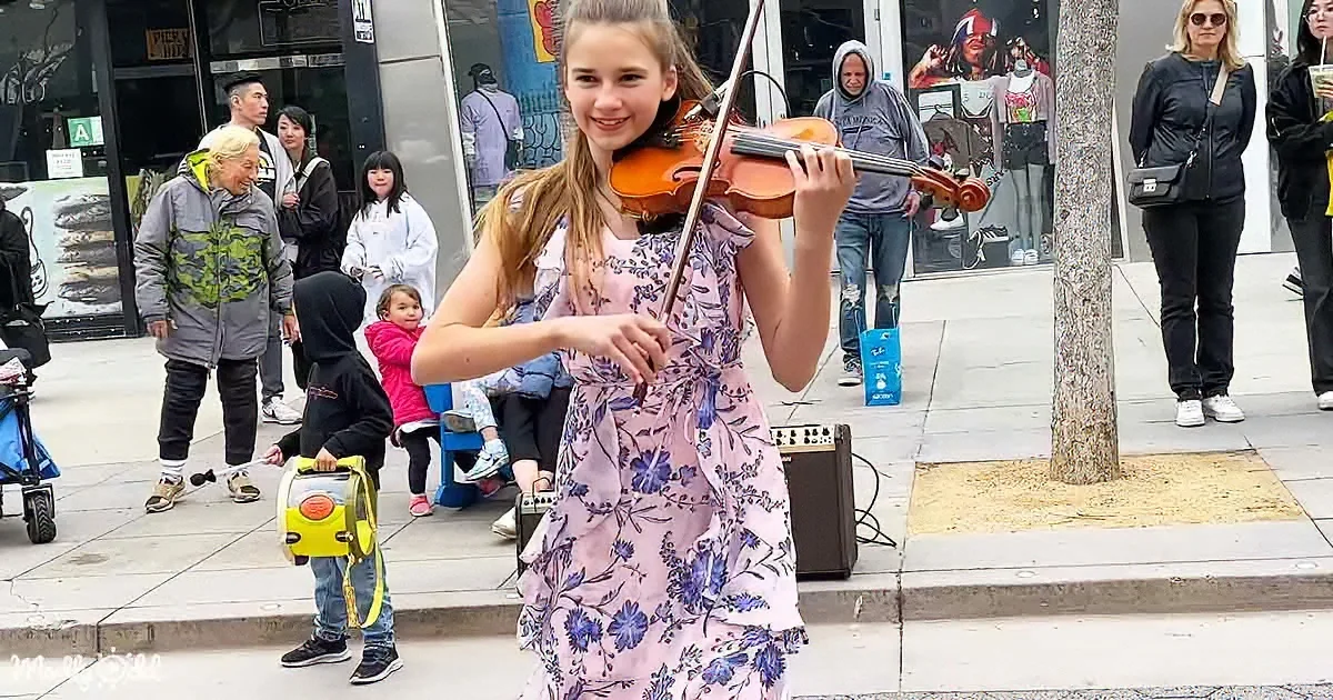 A girl in a floral dress plays violin on a sidewalk, surrounded by onlookers and a child with a yellow case.