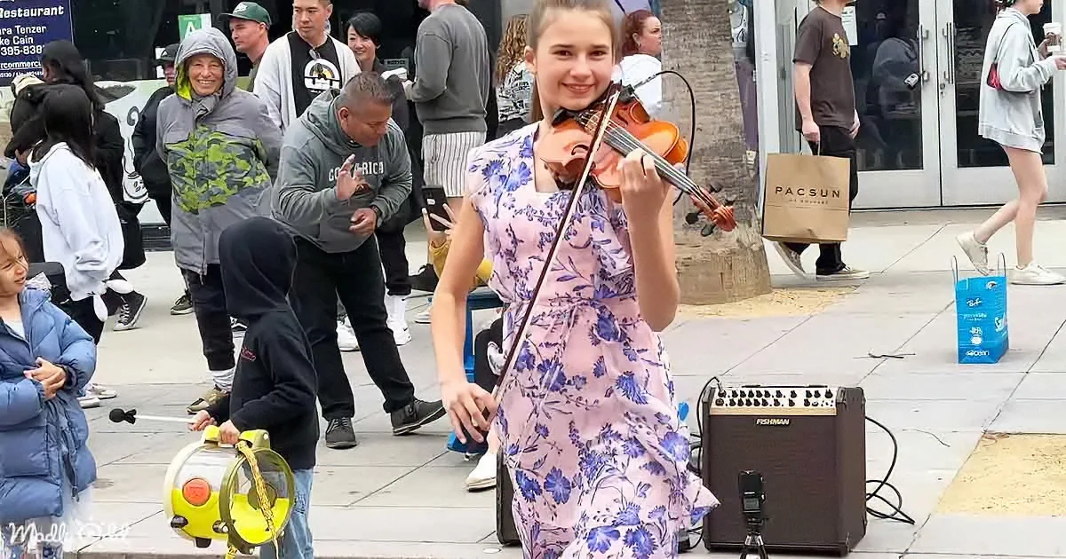 A girl in a floral dress plays violin on a sidewalk, surrounded by people and a PACSUN shopping bag.