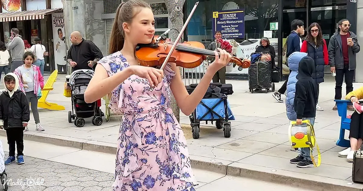 A young girl in a floral dress plays the violin on a busy sidewalk, surrounded by pedestrians and street vendors.