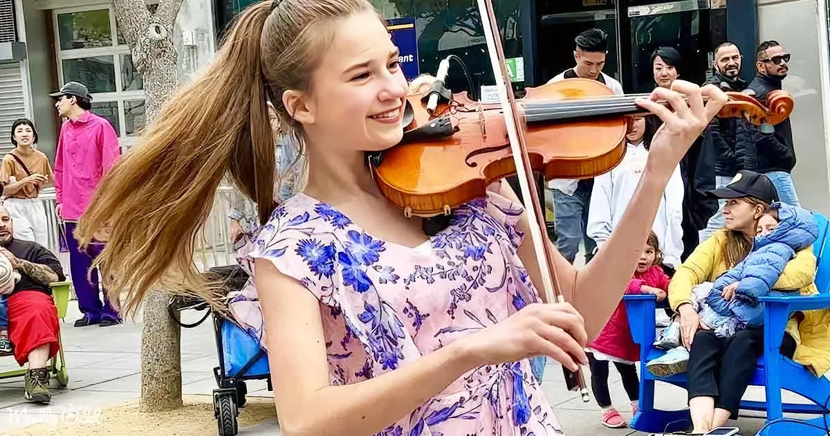 A young woman in a floral dress plays the violin in front of a crowd on a sunny day.
