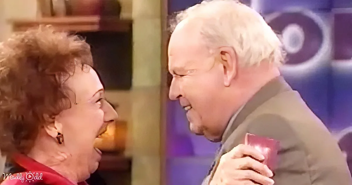 An older couple smiles warmly at each other, holding hands, in a cozy indoor setting.