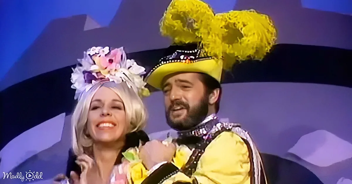 A man in a yellow costume with a large yellow hat and a woman in a floral outfit are smiling and holding hands on stage.