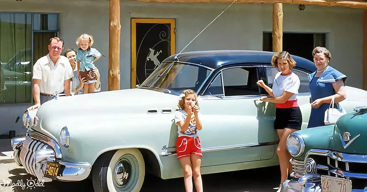 A family poses with a vintage light blue car in front of a building with a horse logo.