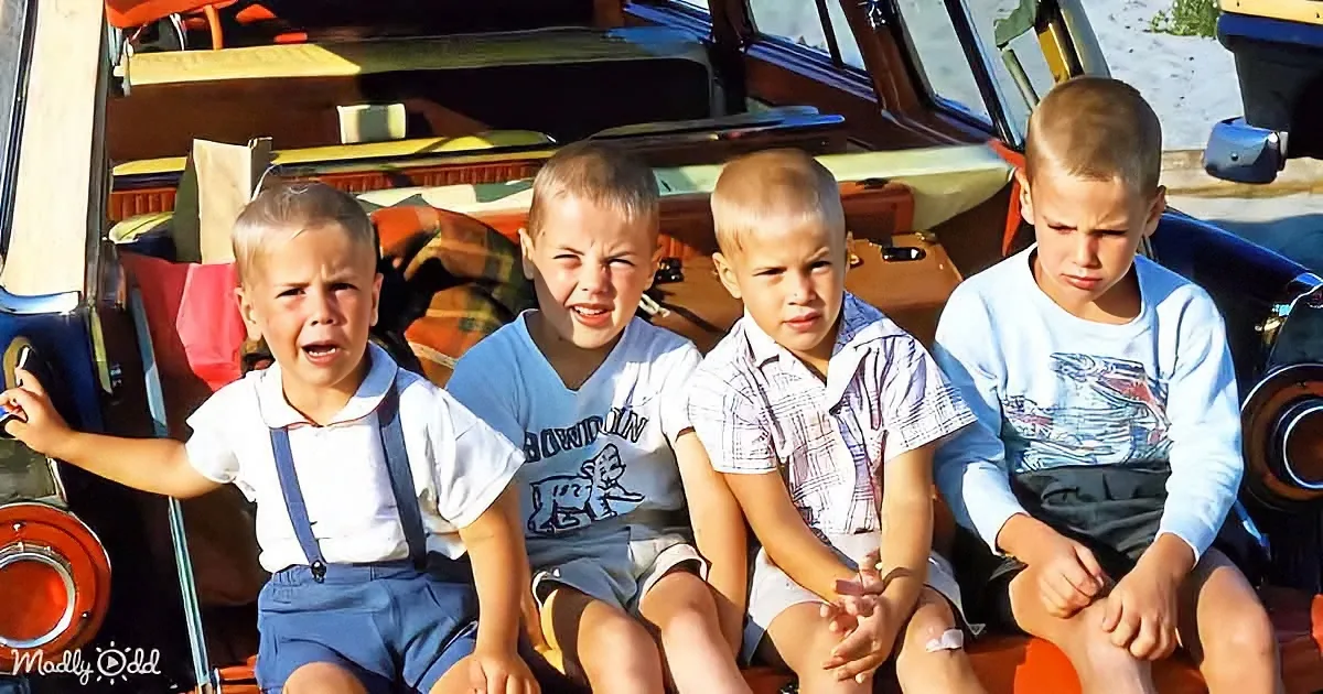 Four young boys sit together, one crying, in front of a vintage car on a sunny day.