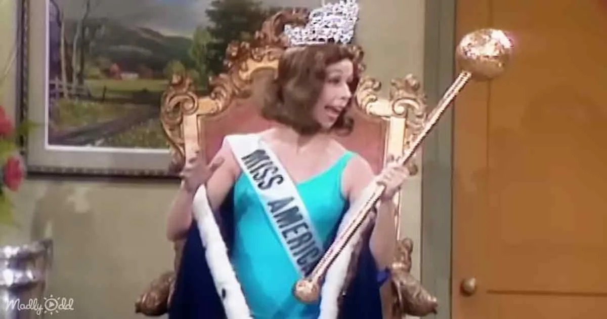 A woman in a blue dress and sash, wearing a crown, holds a scepter, seated on a throne.