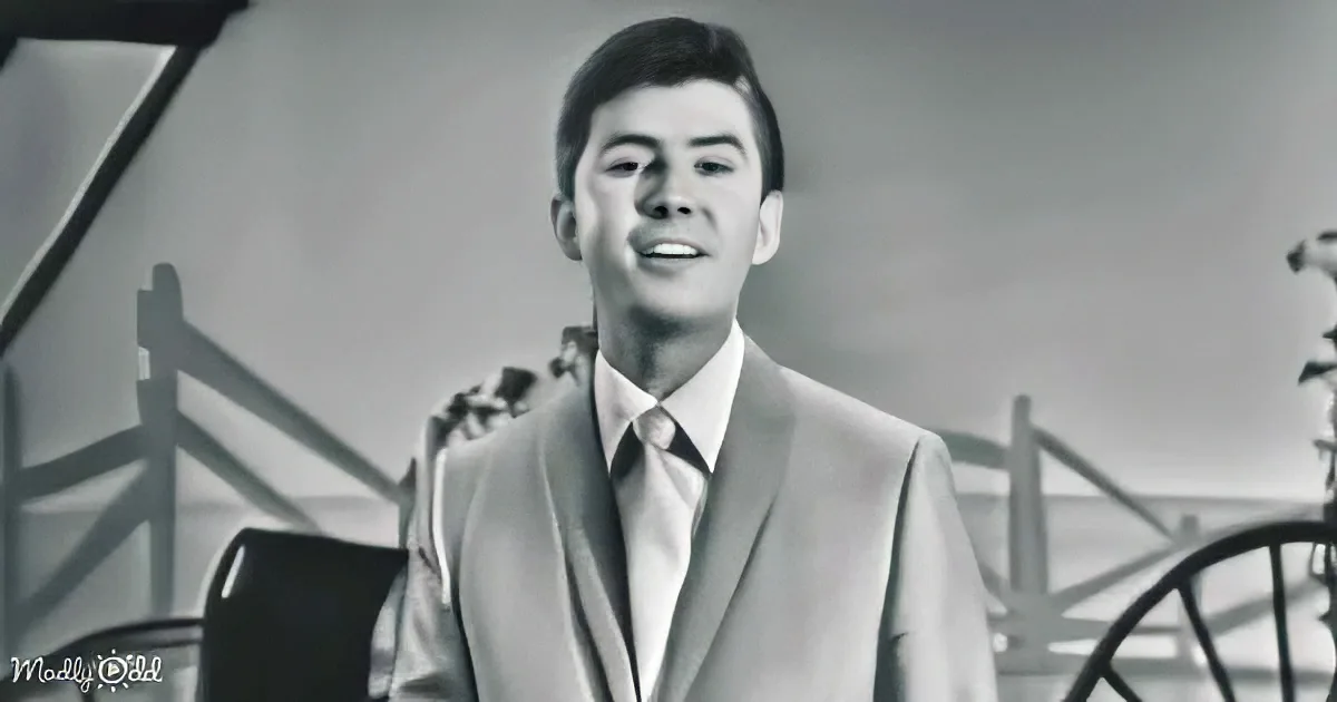 A man in a suit and tie stands in a black-and-white setting, possibly from a film or music video.