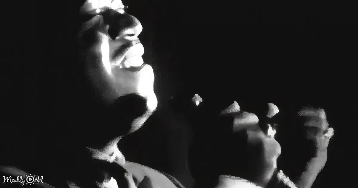 A person in a suit and tie is singing into a microphone, with a crowd in the background.