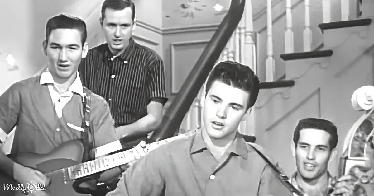 A man in a brown shirt sings while playing guitar, surrounded by other musicians in a vintage setting.