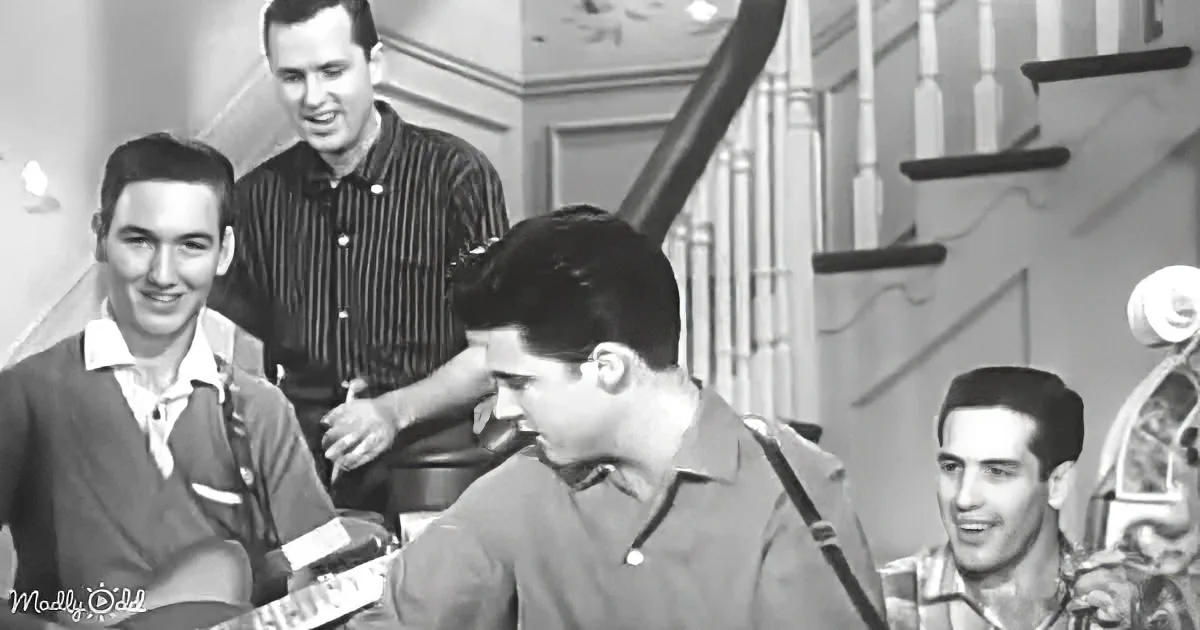 A man in a striped shirt stands behind a seated man playing guitar, with others in the background.