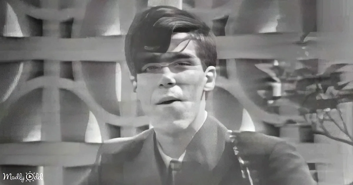 A man with short hair and glasses, wearing a jacket, stands in front of a wooden fence.