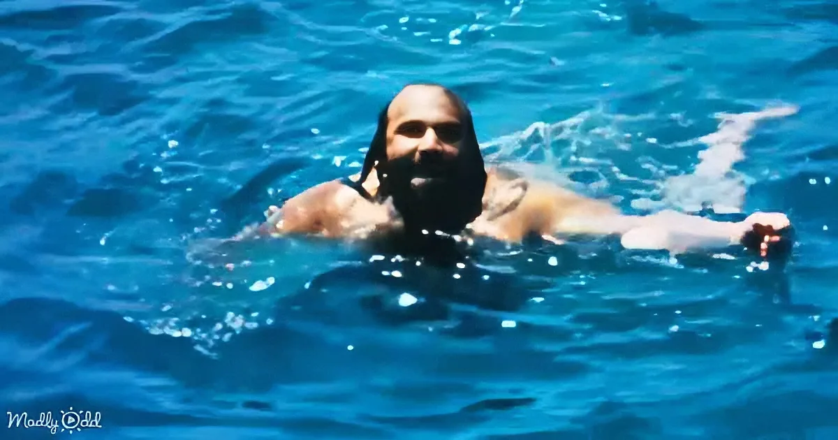 A man with a beard and long hair is swimming in clear blue water, creating ripples around him.