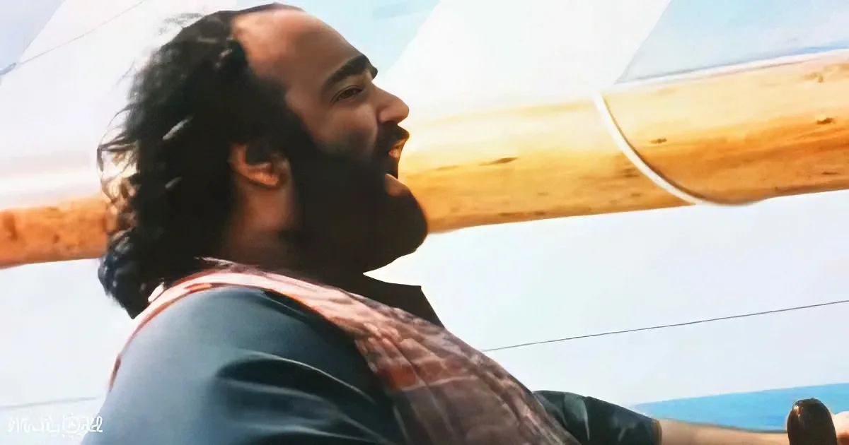 A man with long hair and a beard, wearing a blue shirt and a patterned scarf, is smiling and looking to the side.