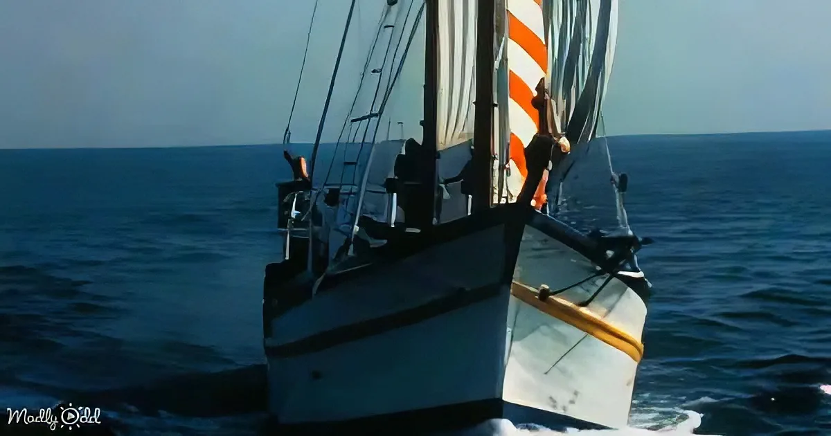A sailboat with striped sails and a yellow rope is sailing on a calm blue ocean under a clear sky.