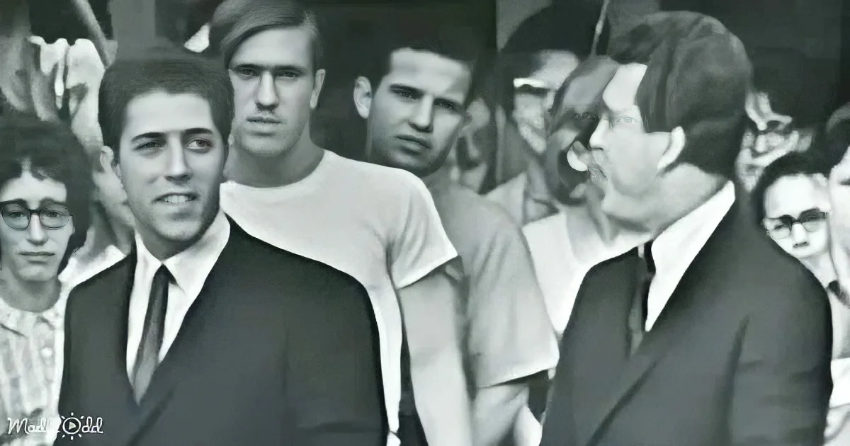 A man in a suit and tie stands among a crowd, looking to the side, with another man in a suit nearby.