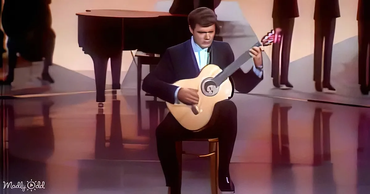 A man in a suit plays a lute on stage, with a piano and dancers in the background.