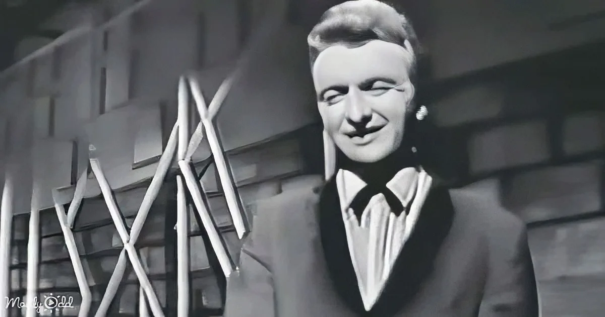 A man in a suit and bow tie smiles in a black-and-white photo with a building in the background.