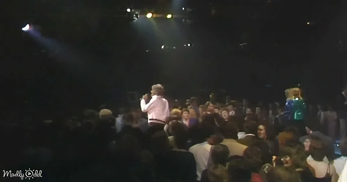 A person in a white shirt stands on stage, surrounded by a crowd, with stage lights above.