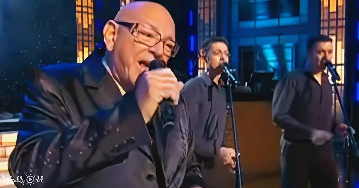 A man in a dark suit sings into a microphone on stage, with two other musicians behind him.