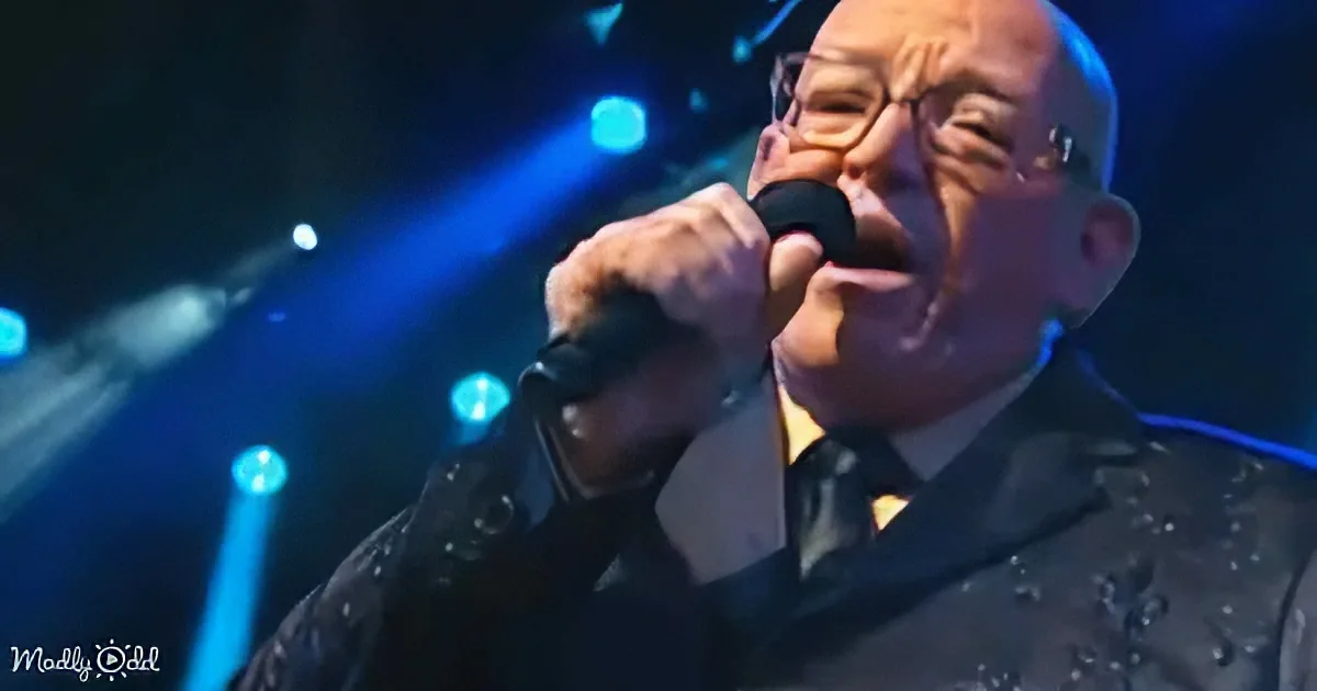 A man in a suit and tie sings into a microphone on stage with blue lighting.