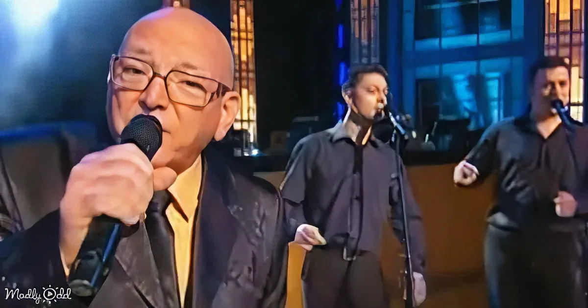 An older man with glasses sings into a microphone, accompanied by two other musicians on stage.