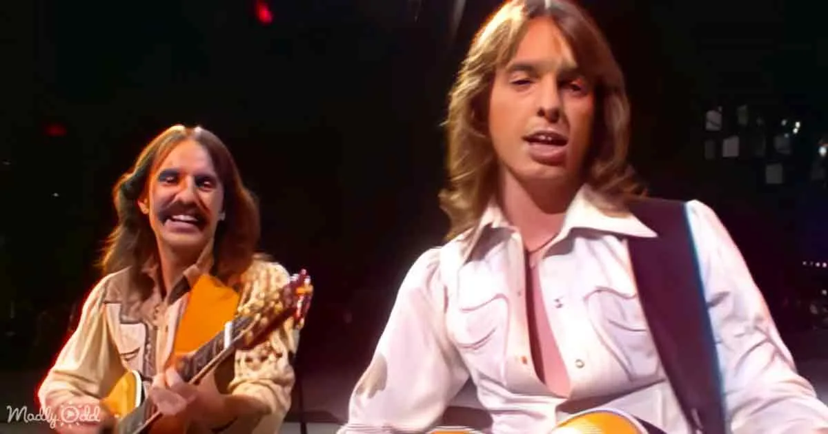 A man with long hair and a white shirt is singing, another man with long hair and a patterned shirt is playing guitar.
