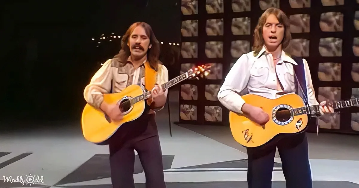 Two musicians with long hair and mustaches play acoustic guitars on stage, wearing 70s-style outfits.