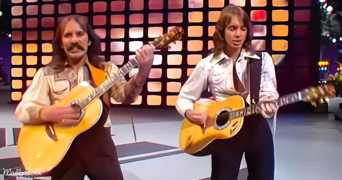 Two musicians with long hair play guitars on a stage with a colorful, grid-patterned backdrop.