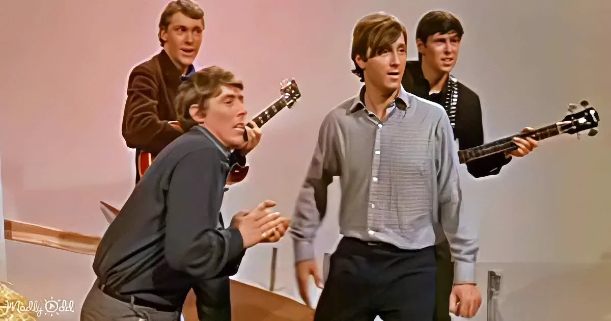 A man in a gray shirt and dark pants is singing and clapping, surrounded by three other men playing guitars.