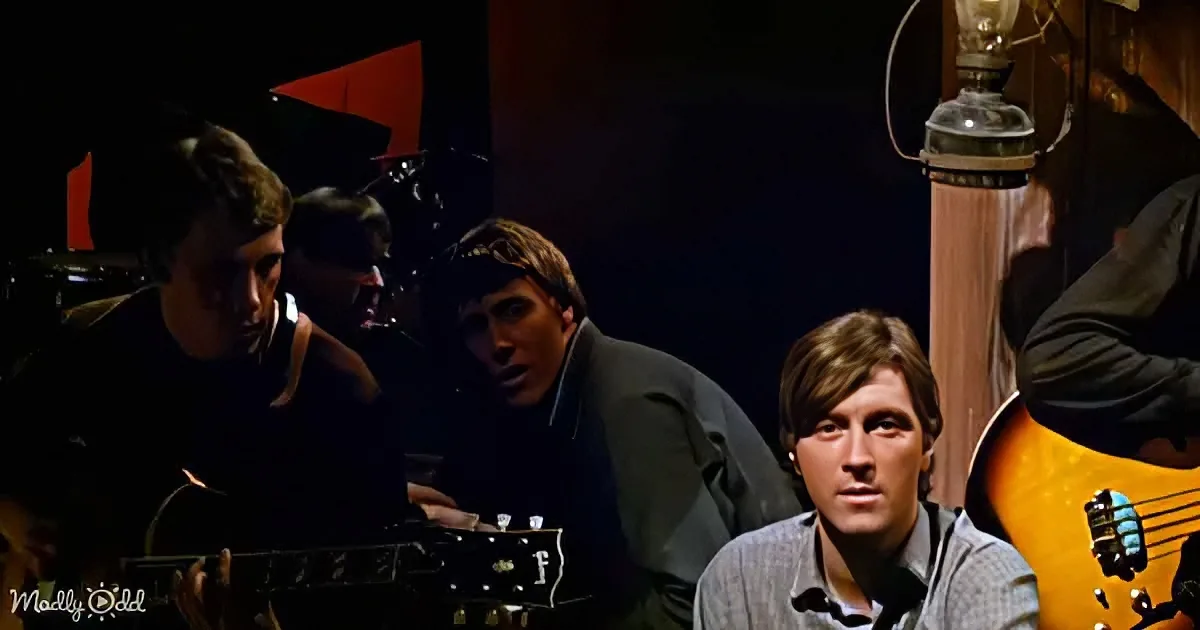 A man in a striped shirt sits in front of a guitar, surrounded by other musicians in a dimly lit room.