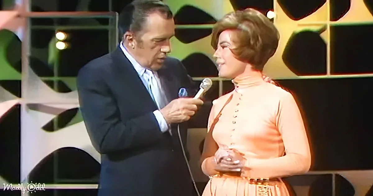 A man in a suit holds a microphone to a woman in a peach dress on stage.