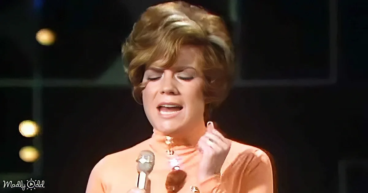 A woman in an orange dress sings into a microphone on stage, with a blurred background and warm lighting.