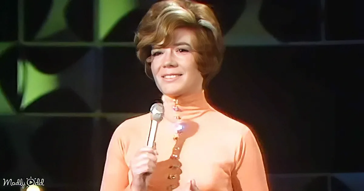 A woman in a peach dress holds a microphone, smiling on stage with a green geometric background.