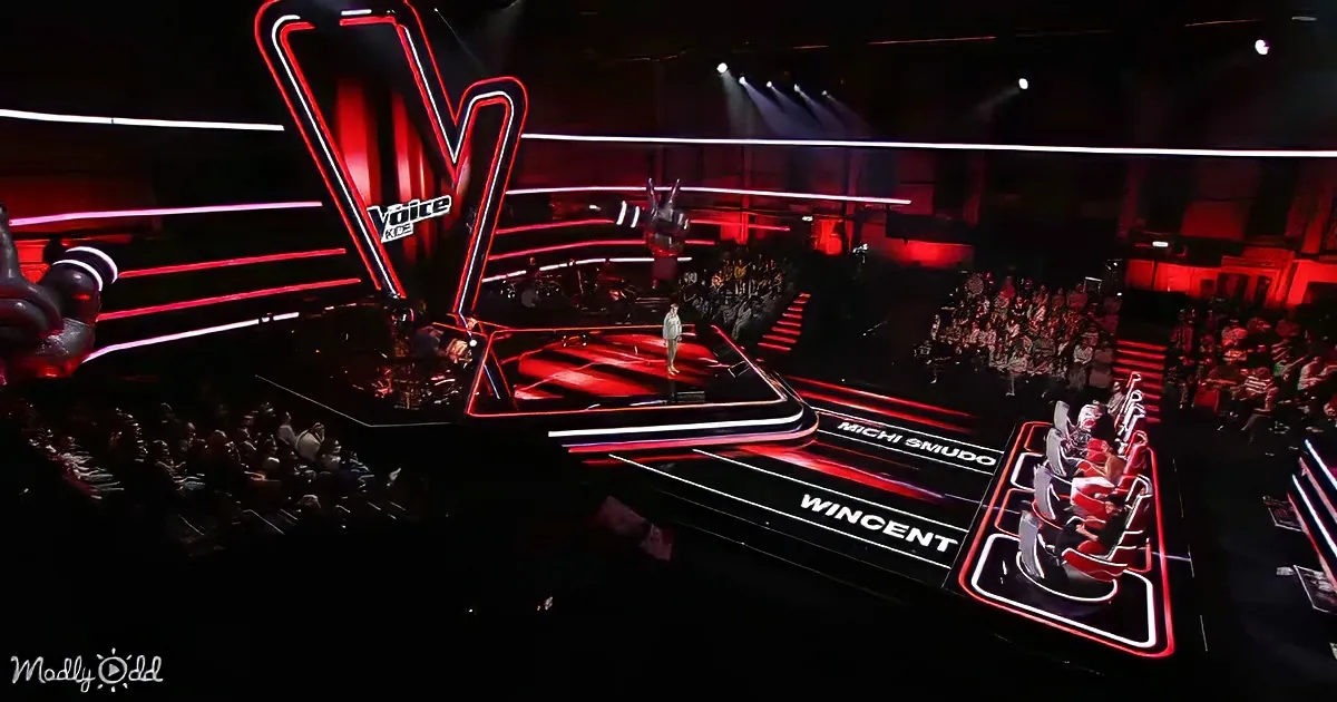 A contestant stands on a stage with red and white lighting, surrounded by judges' chairs and an audience.