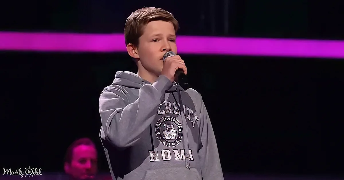 A young boy in a grey hoodie sings into a microphone on stage, with a purple light in the background.