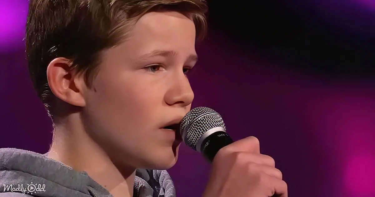 A young boy sings into a microphone on a stage with a purple background.