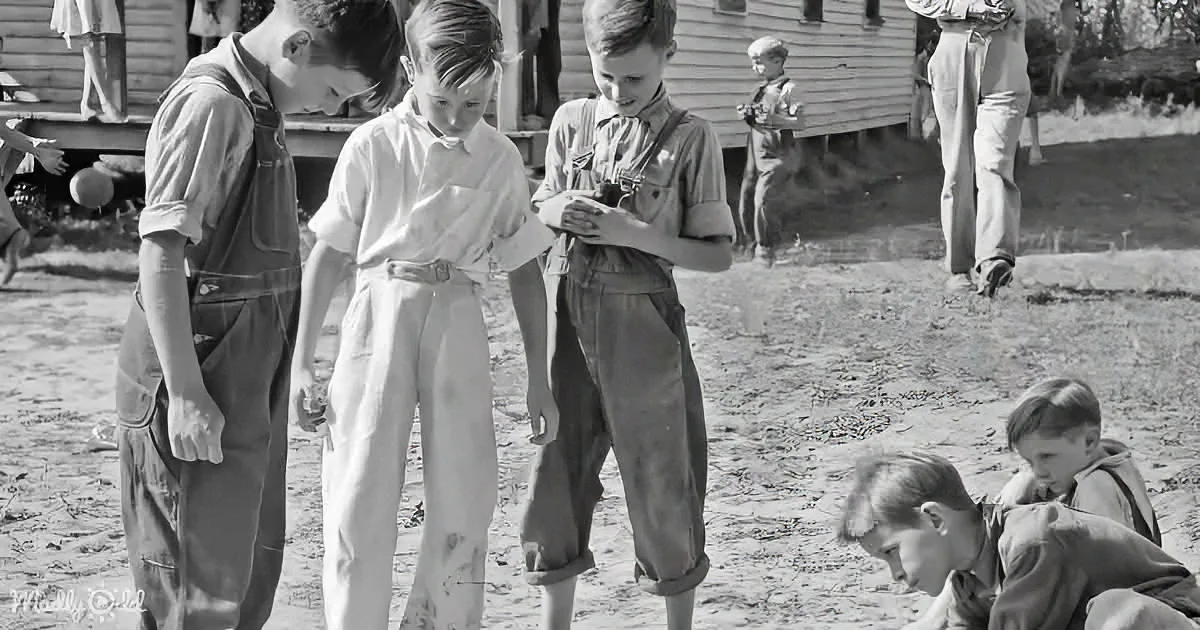 Boys in overalls gather around, one holding a small object, in a rural outdoor setting.