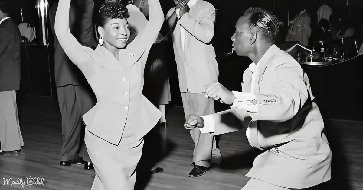 A woman in a suit dances joyfully with a man in a suit, surrounded by other dancers in a lively social setting.
