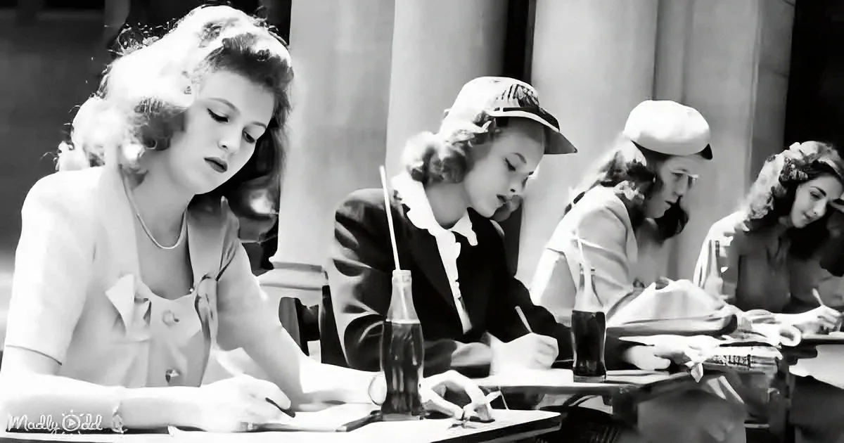 A woman in a white blouse is writing at a table with a bottle of soda, surrounded by other students in a classroom setting.