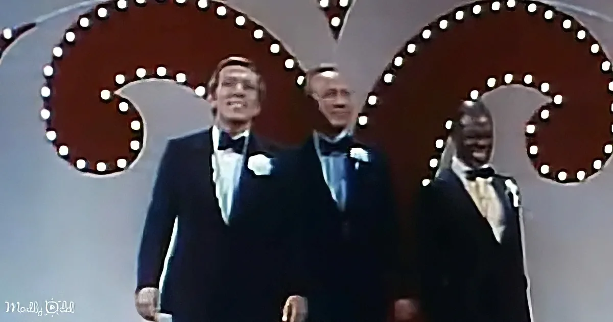 Three men in tuxedos stand on stage, smiling, with a large illuminated "G" in the background.