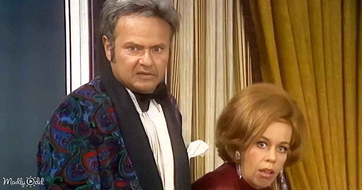 A man in a colorful paisley jacket and a woman in a red dress are standing in front of a curtain.
