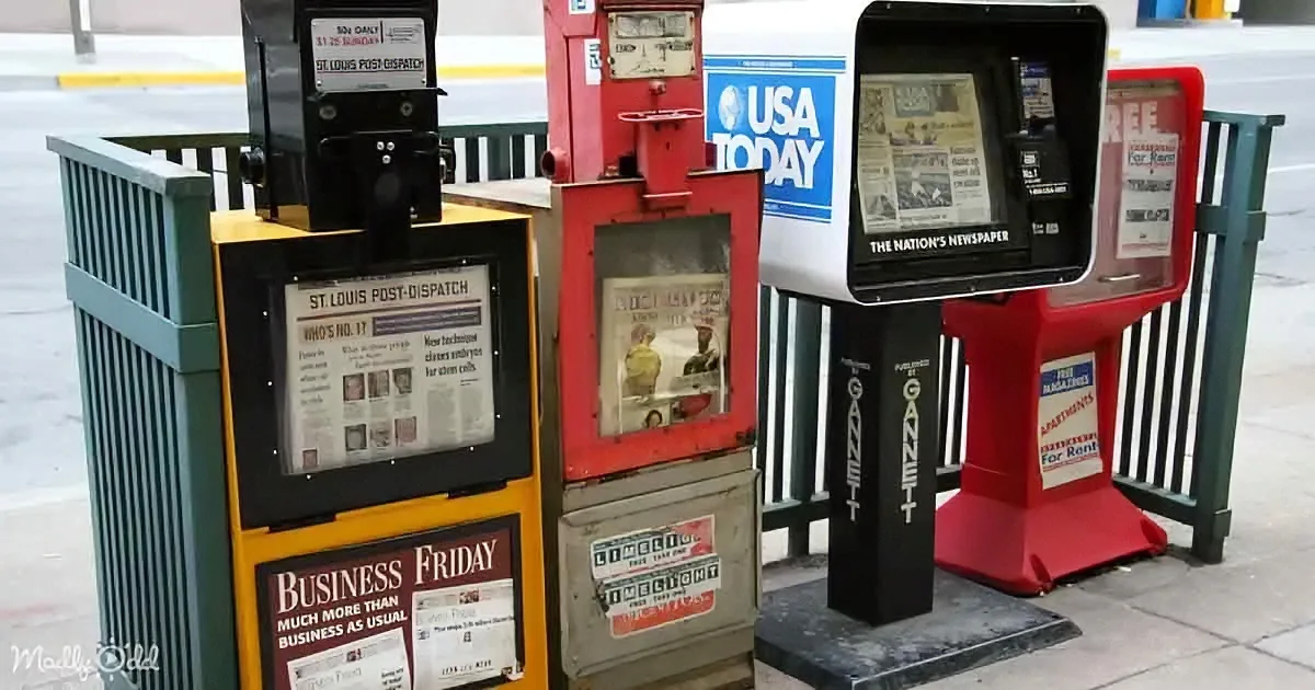 Multiple vintage newspaper vending machines display various local and national newspapers on a city sidewalk.