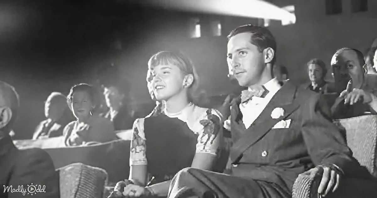 A man and woman in formal attire sit together, smiling, in a dimly lit theater audience.