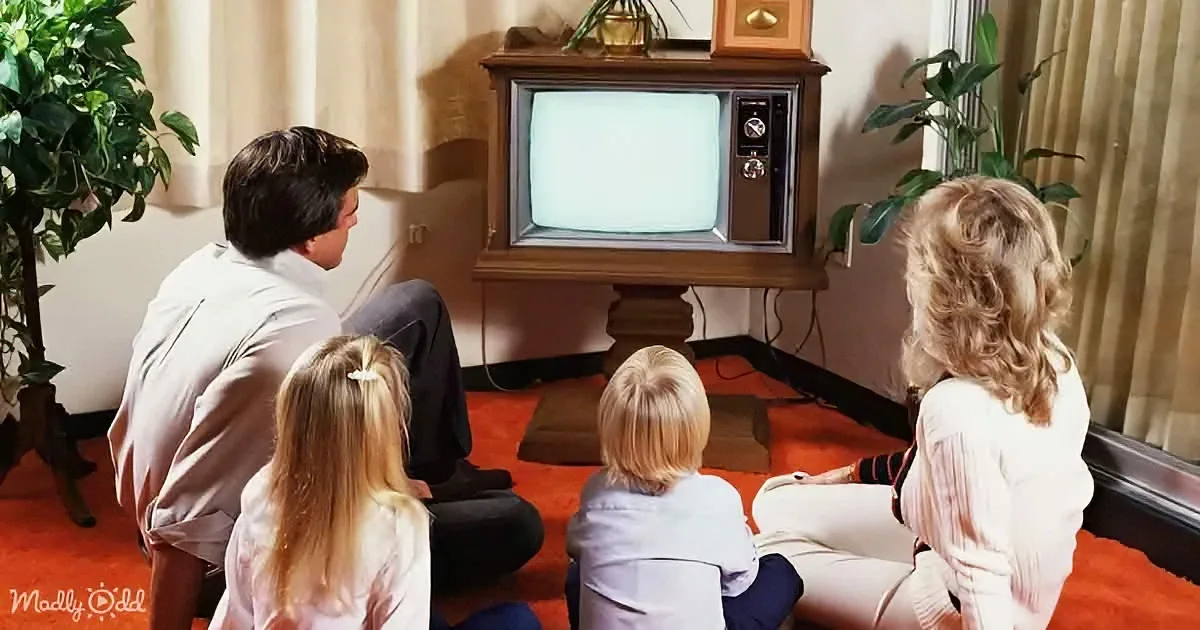 A family of four sits on the floor watching an old-fashioned TV in a cozy living room.