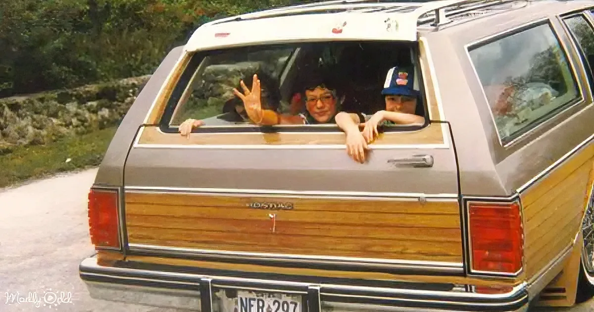 A vintage station wagon with wood paneling has two children waving from the back window.