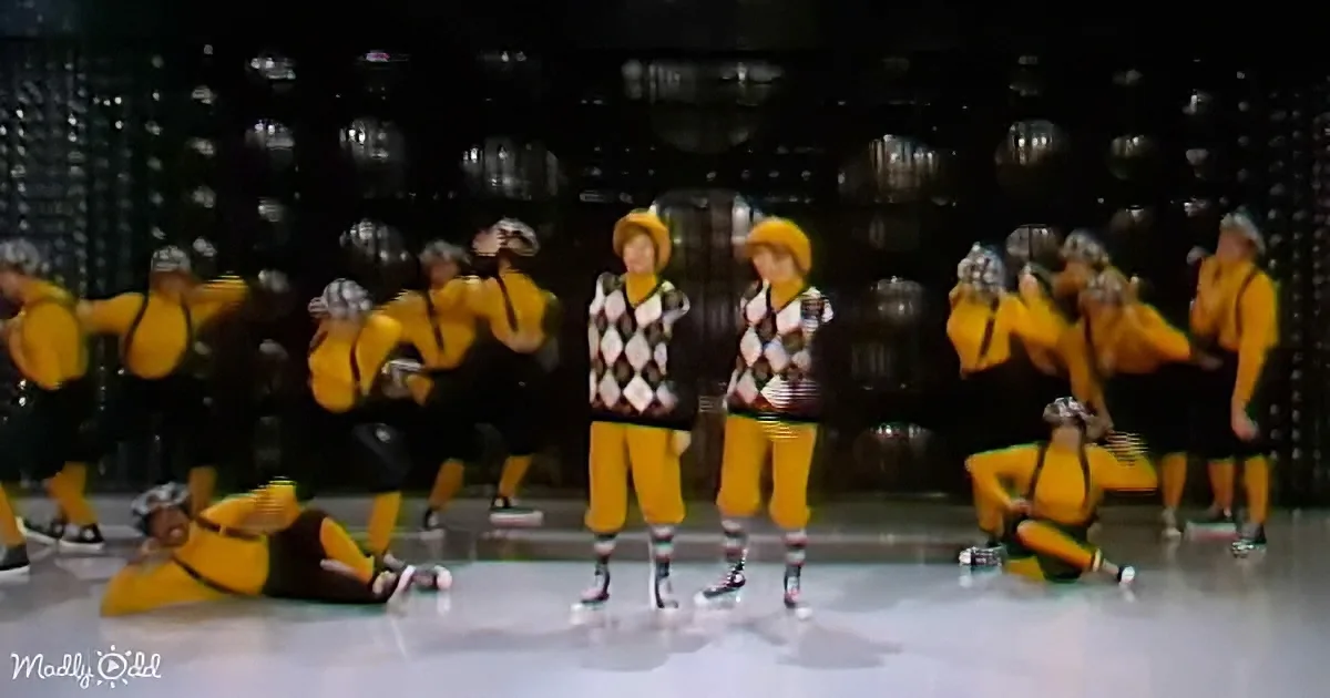 Two dancers in argyle sweaters and yellow pants stand center stage, surrounded by performers in yellow outfits.