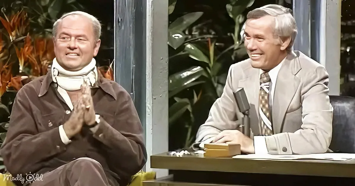 Two men are seated, one in a suit smiling, the other in a brown jacket with hands clasped, both appear to be in a studio...