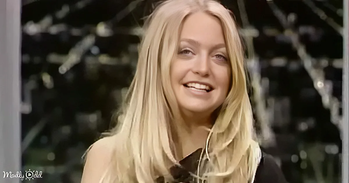 A smiling person with long blonde hair, wearing a dark top, stands in front of a blurred background with visible structures.