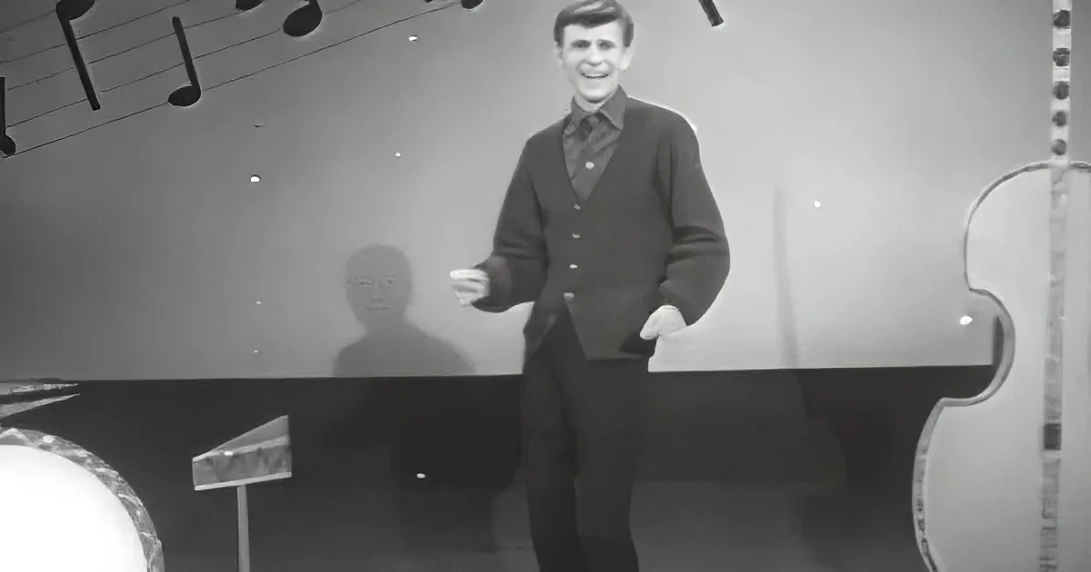 A man in a sweater and tie stands in front of a piano, smiling, with musical notes above him.