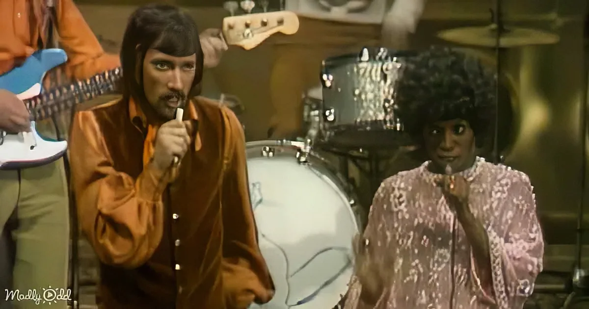 A man in a brown shirt holds a microphone, singing, with a woman in a patterned dress and afro in the background.